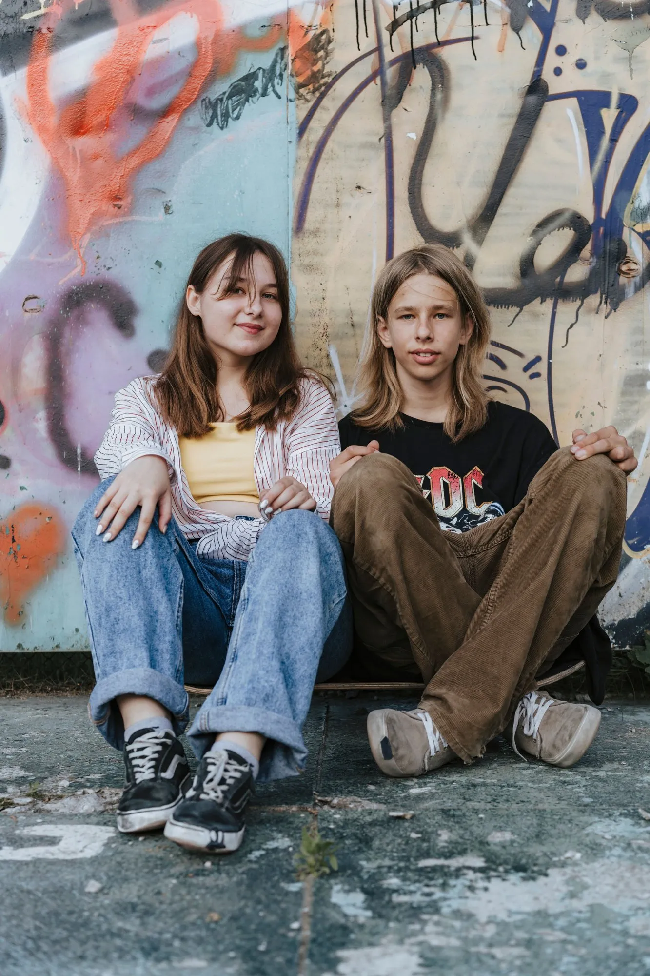 Ein Mädchen und ein Bursch sitzen nebeneinander vor einer Graffitiwand.