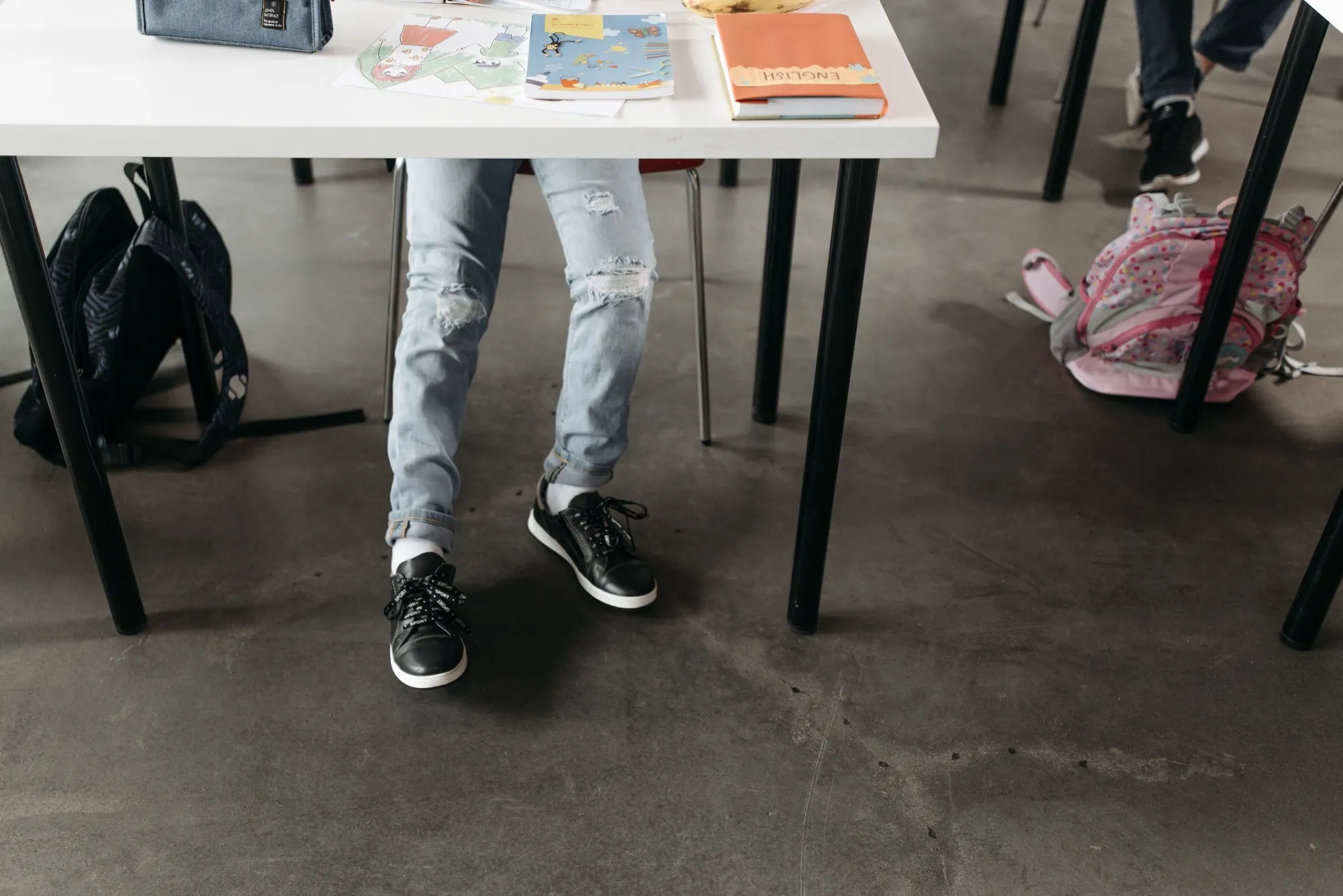 Die Beine eines Kindes in Jeans und Turnschuhen unter einem Schultisch, daneben Schultaschen am Boden.